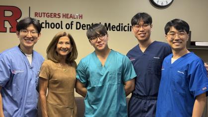 A group of five individuals standing together in front of a beige wall with the Rutgers Health School of Dental Medicine logo. Four people are wearing medical scrubs in shades of blue and teal, while one person is dressed in a brown short-sleeve dress. A clock is visible on the wall above them.