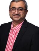 Man with short gray and black hair, glasses, and a mustache, wearing a black blazer over a red and white checkered shirt, smiling at the camera against a plain white background.