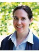 Portrait of a woman with light skin, blue eyes, and dark hair pulled back, smiling at the camera. She is wearing a white collared shirt under a dark sweater, with a blurred background of green foliage.