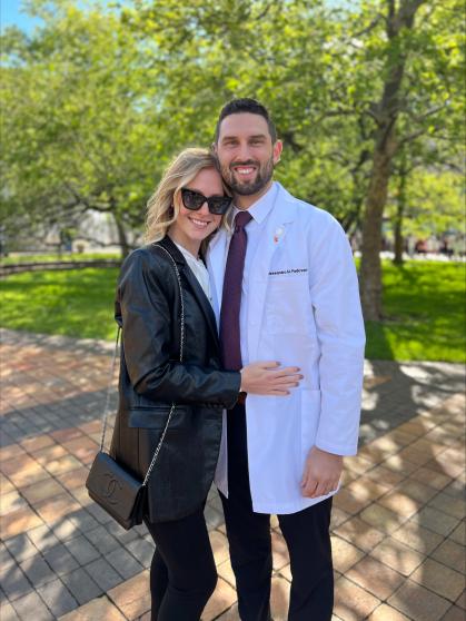 "A person wearing a white medical coat with a name tag that reads Alessandro Padovani stands next to another person dressed in a black leather jacket and carrying a black handbag. They are standing outdoors on a sunny day, with green trees and grass in the background. 