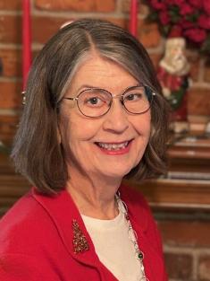 A smiling woman with short brown and gray hair, wearing glasses, a red blazer, and a Christmas-themed brooch. She is in front of a brick fireplace decorated with red candles, flowers, and holiday figurines.