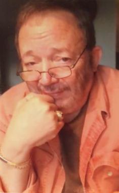 A person wearing a light orange button-up shirt over a dark inner garment, resting their chin on their hand. The hand is adorned with a gold ring and a bracelet. The background is softly lit and out of focus.